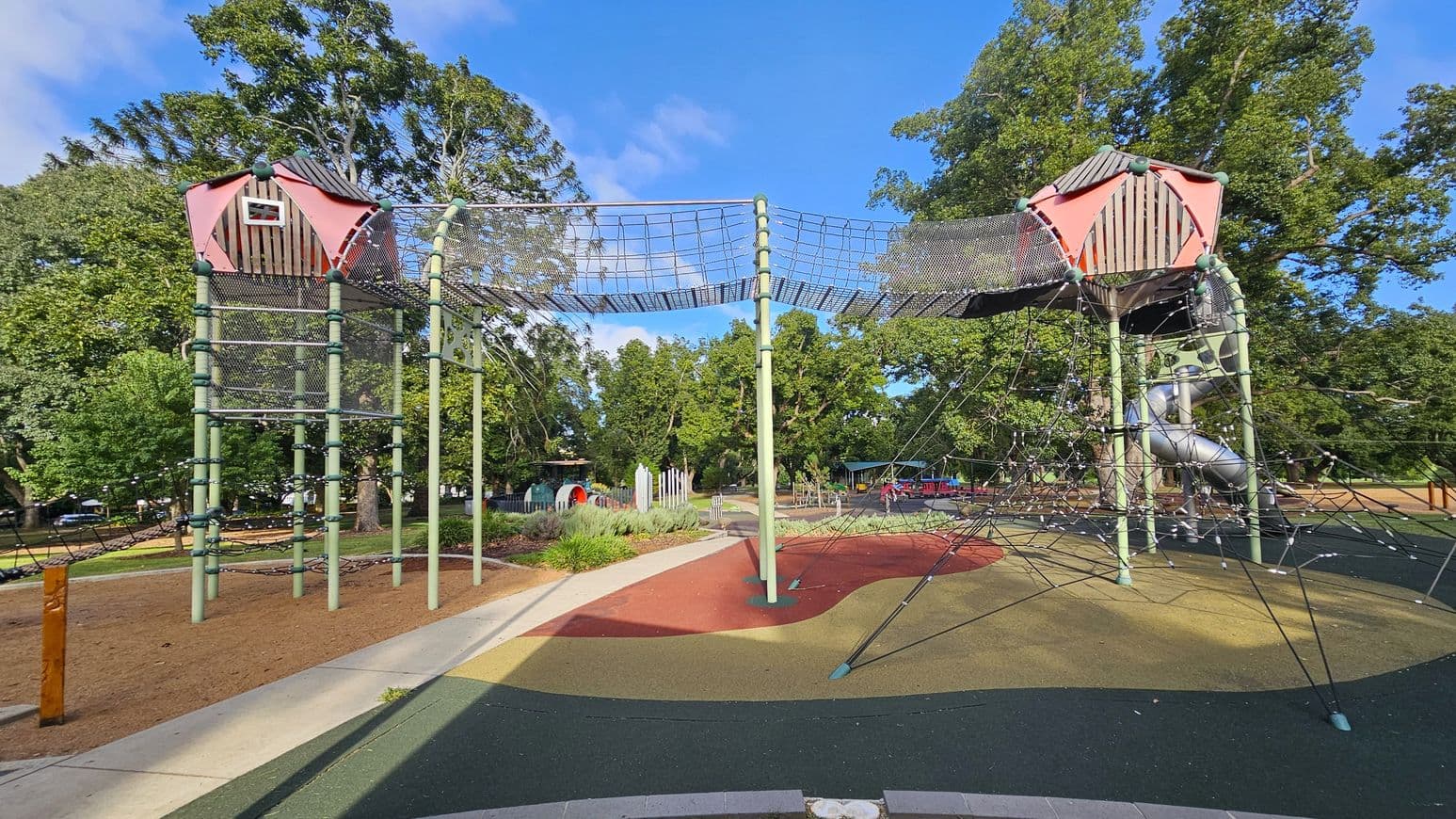 A playground with two elevated structures connected by a net bridge. Each structure has a red and grey roof resembling a small house. The left structure has a climbing net, while the right one features a spiral slide. The ground is covered with colourful rubber surfacing in green, red, and yellow. Surrounding the playground are trees and additional play equipment in the background. The area is bordered by a concrete path.