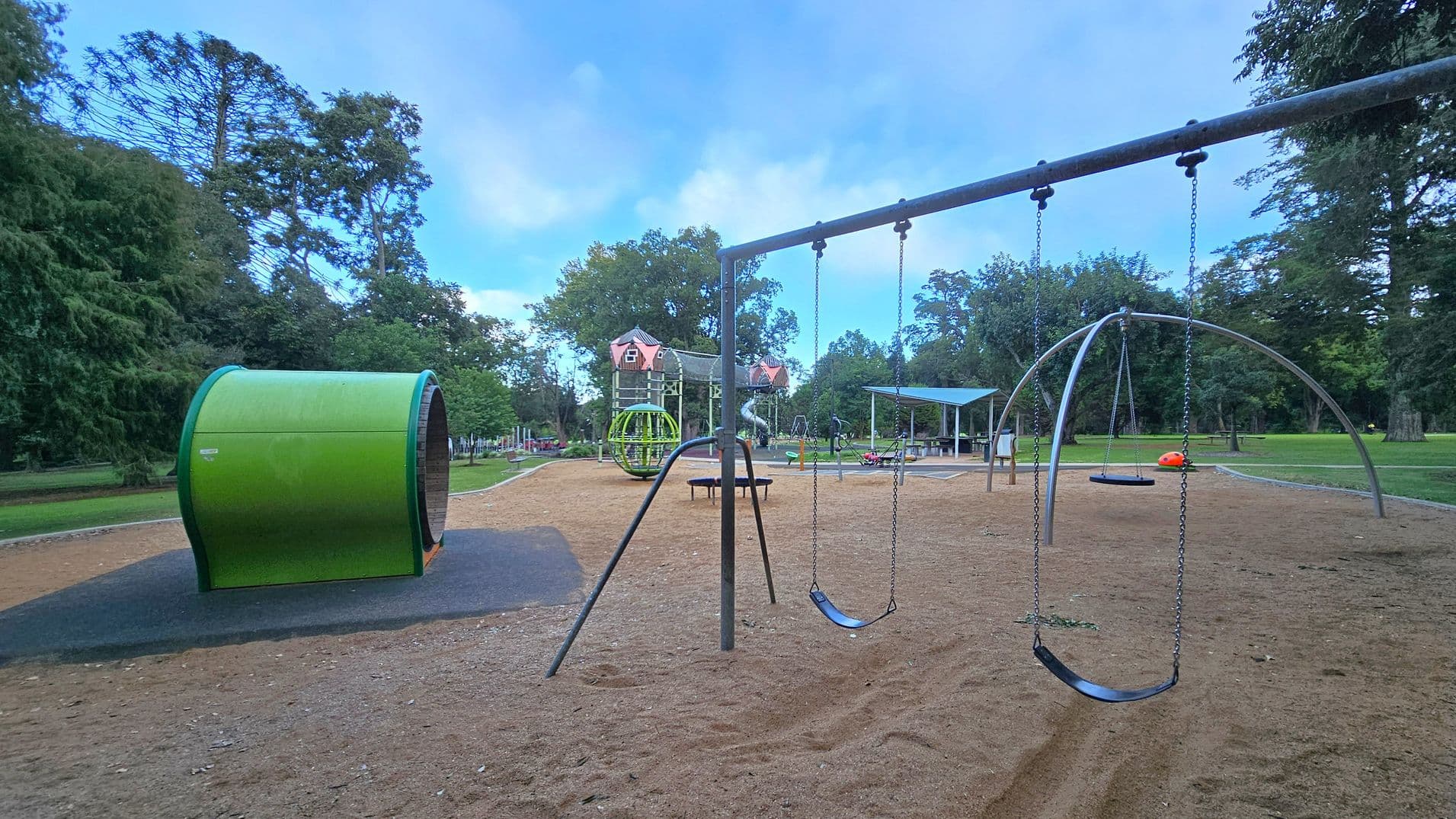 A playground with various equipment. On the left, there's a large green cylindrical structure. In the centre, a swing set with two swings hangs from a metal frame. Behind it, a climbing structure with red-roofed towers is visible. To the right, a covered seating area and a small orange play element are present. The ground is covered with wood chips, and the area is surrounded by trees and grass.