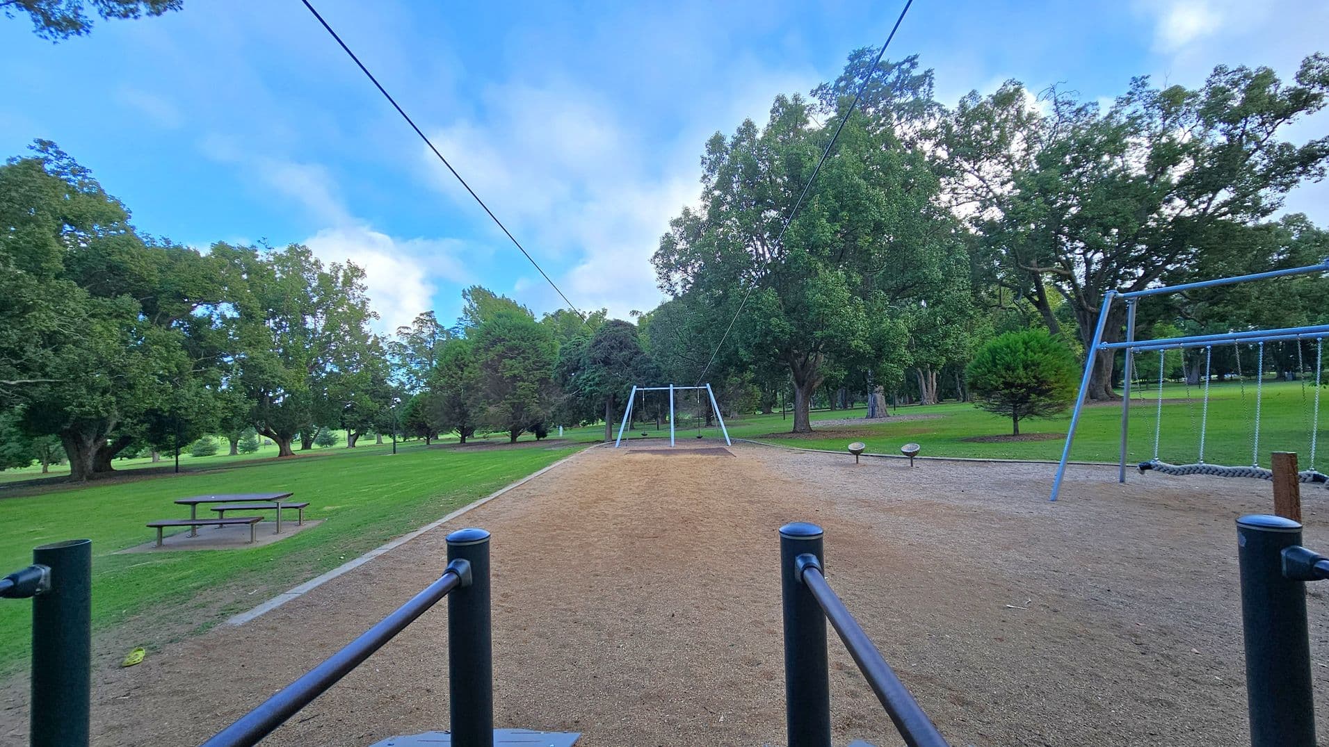 A park scene with a zip line in the centre, stretching across a sandy area. On the right, there's a swing set with a netted swing. To the left, a picnic table sits on a grassy area. The background is filled with large, leafy trees. The ground is a mix of grass and sand, and the sky is visible above.