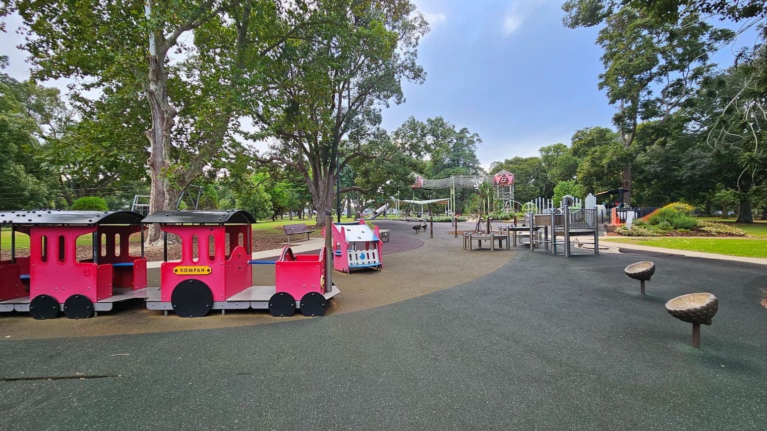 A playground with a red toy train structure on the left, featuring a locomotive and a carriage. In the centre, there is a small white and pink play structure. The right side has various playground equipment, including climbing frames and swings. The ground is covered with a mix of black and brown rubber safety flooring. Large trees with green foliage surround the area, providing shade. A bench is visible in the background under the trees.