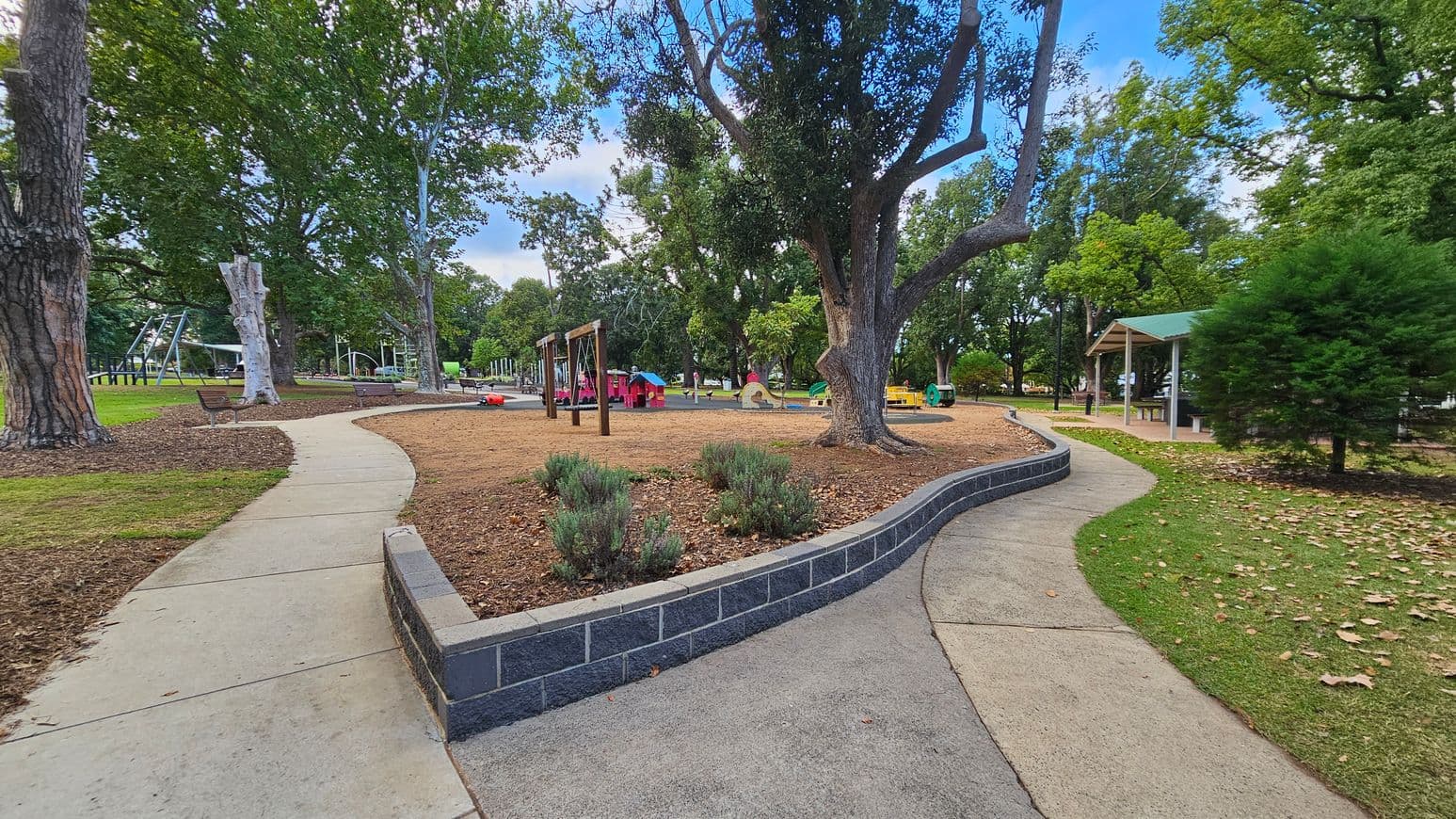 A park with a winding concrete path bordered by grass and mulch. On the left, there are large trees and benches. In the centre, a playground area features colourful play structures and swings. To the right, a covered picnic area with a green roof is visible, along with more trees and a small bush. The ground is a mix of grass, mulch, and paved surfaces.