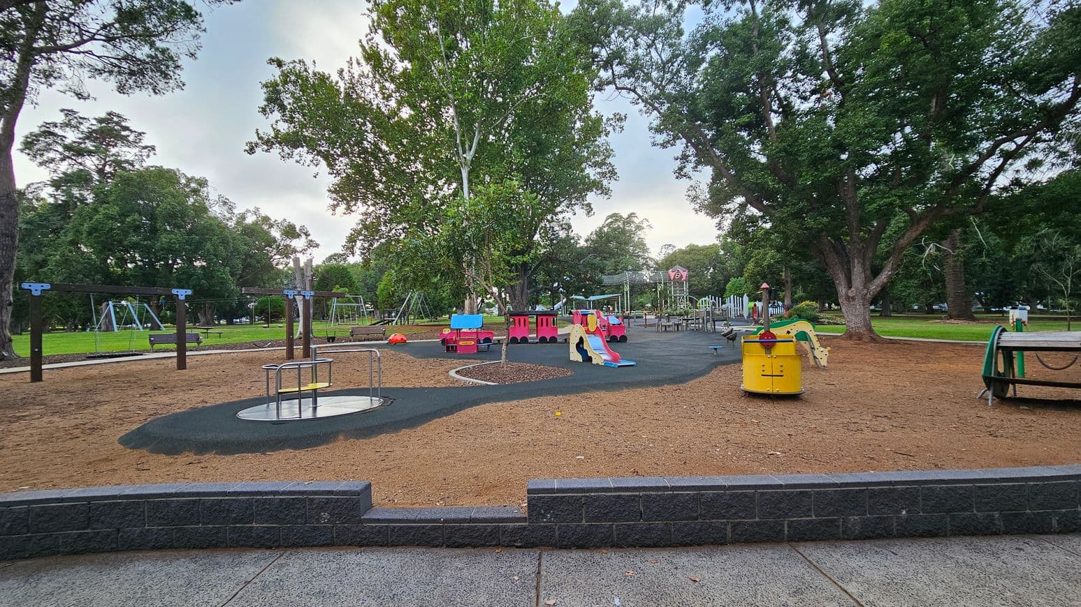 A playground with various equipment on a sandy surface. In the centre, there's a colourful play structure resembling a train with a slide. To the left, swings are visible among green grass and trees. On the right, a yellow roundabout and a spring rider shaped like a horse are present. Large trees surround the area, providing shade. The foreground has a paved path with a low brick wall bordering the playground.