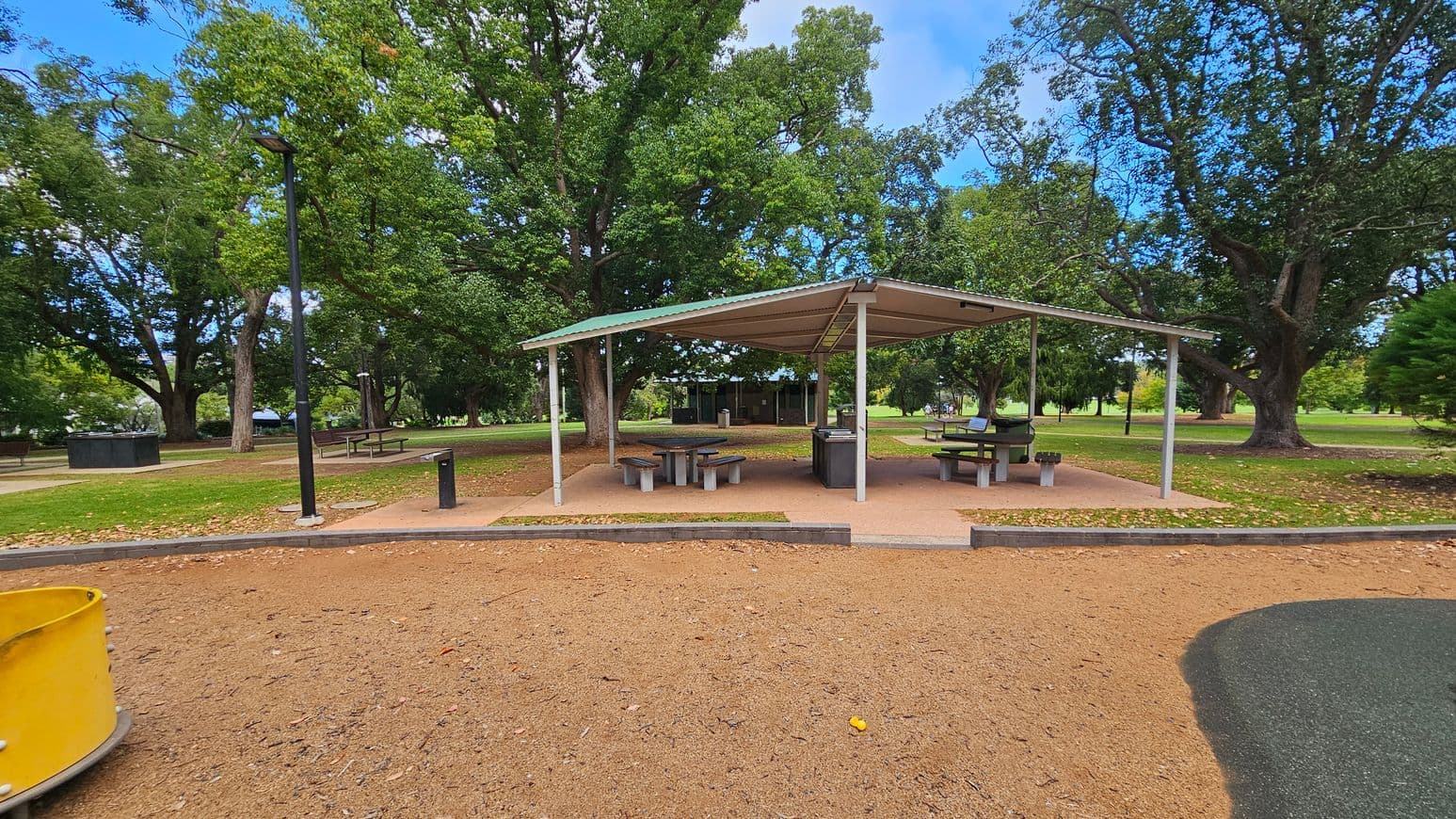 A park scene with a covered picnic area in the centre, featuring several tables and benches. The ground is a mix of grass and dirt, with a paved path leading to the shelter. A yellow play structure is partially visible on the left. Tall trees with dense green foliage surround the area, providing shade. A black lamp post stands near the shelter. In the background, more trees and open grassy spaces are visible.