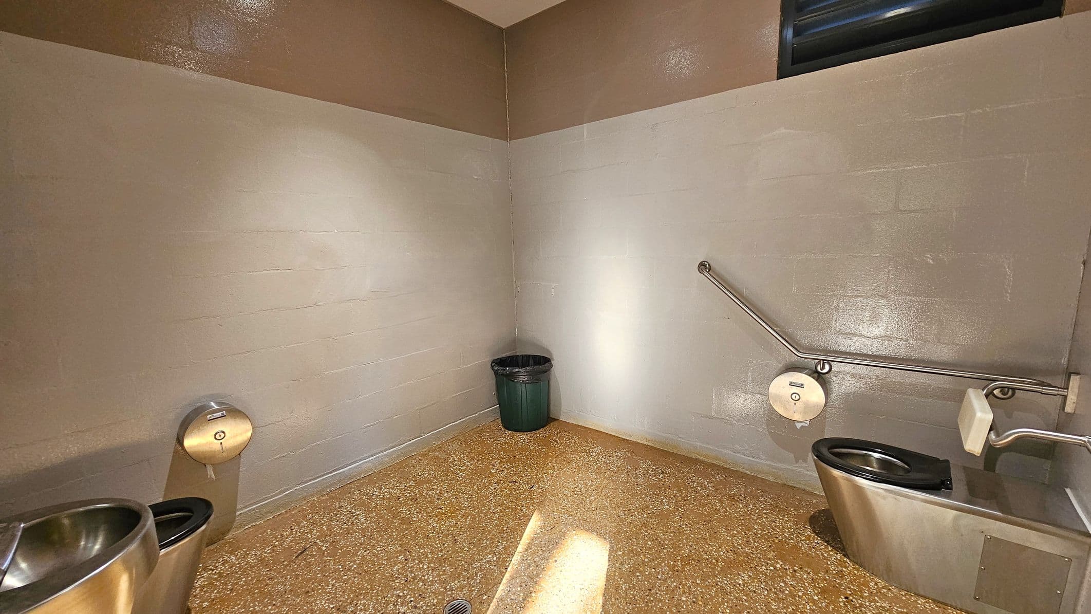A public restroom with a stainless steel toilet on the right, equipped with a black seat and a metal grab bar above it. A toilet paper dispenser is mounted on the wall. On the left, a stainless steel sink is partially visible. In the corner, a green rubbish bin is placed against the light-coloured tiled walls. The floor is speckled with a terrazzo pattern, and a small drain is visible. The ceiling is plain with a small vent near the top of the wall.