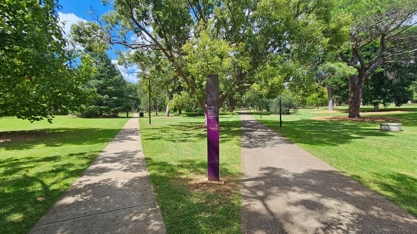 A park scene with a central path splitting into two, bordered by lush green grass and trees. In the middle, a purple signpost provides directions. The left path is shaded by a large tree, while the right path is lined with smaller trees and a bench. The background features more trees and open grassy areas. Shadows of the trees are cast on the paths.
