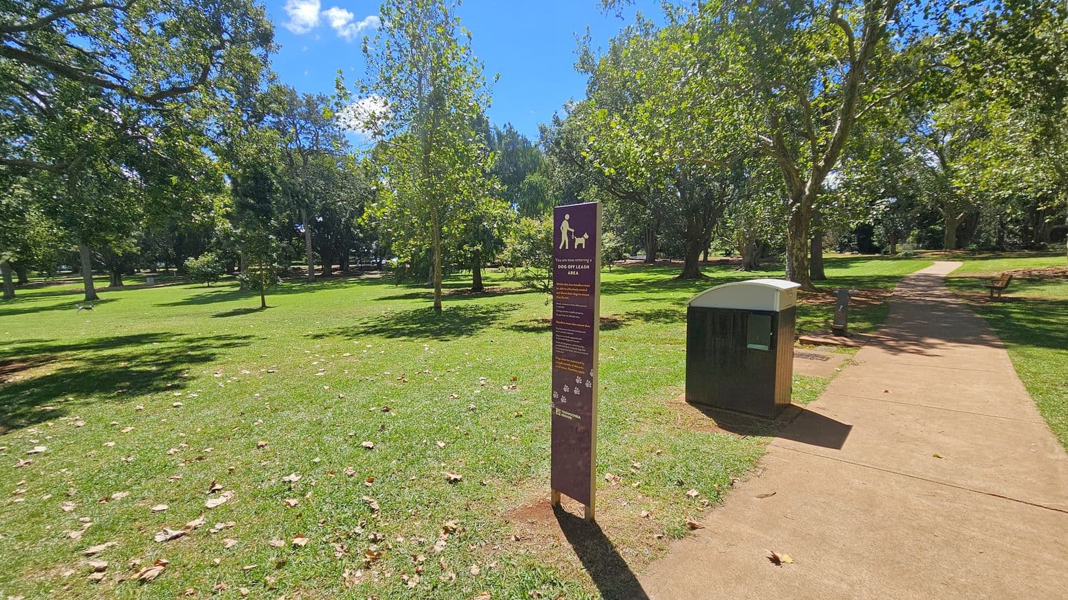 A park scene with a grassy area and scattered trees. In the centre, a sign indicates a dog off-leash area. To the right, a paved path runs alongside a rubbish bin. Benches are visible further along the path. The ground is covered with green grass and some fallen leaves.
