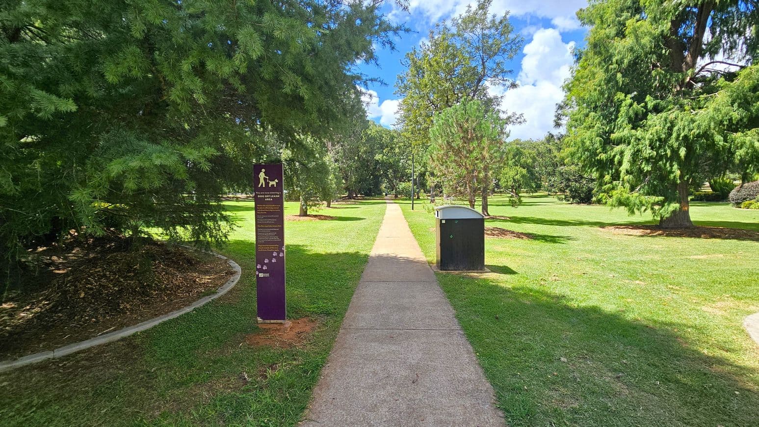 A park scene with a concrete path running through the centre, flanked by green grass and trees. On the left, a purple sign indicates a dog off-leash area. On the right, a black bin is positioned near the path. The background features more trees and open grassy areas under a partly cloudy sky.