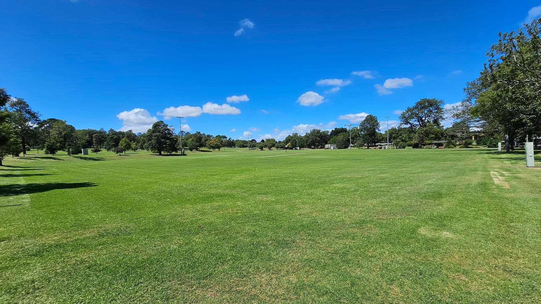 A vast, open grassy field stretches across the foreground, with a few patches of brown. On the left and right, trees line the perimeter, casting shadows on the grass. In the distance, more trees and some structures are visible, along with several tall light poles. The sky is clear with a few scattered clouds.