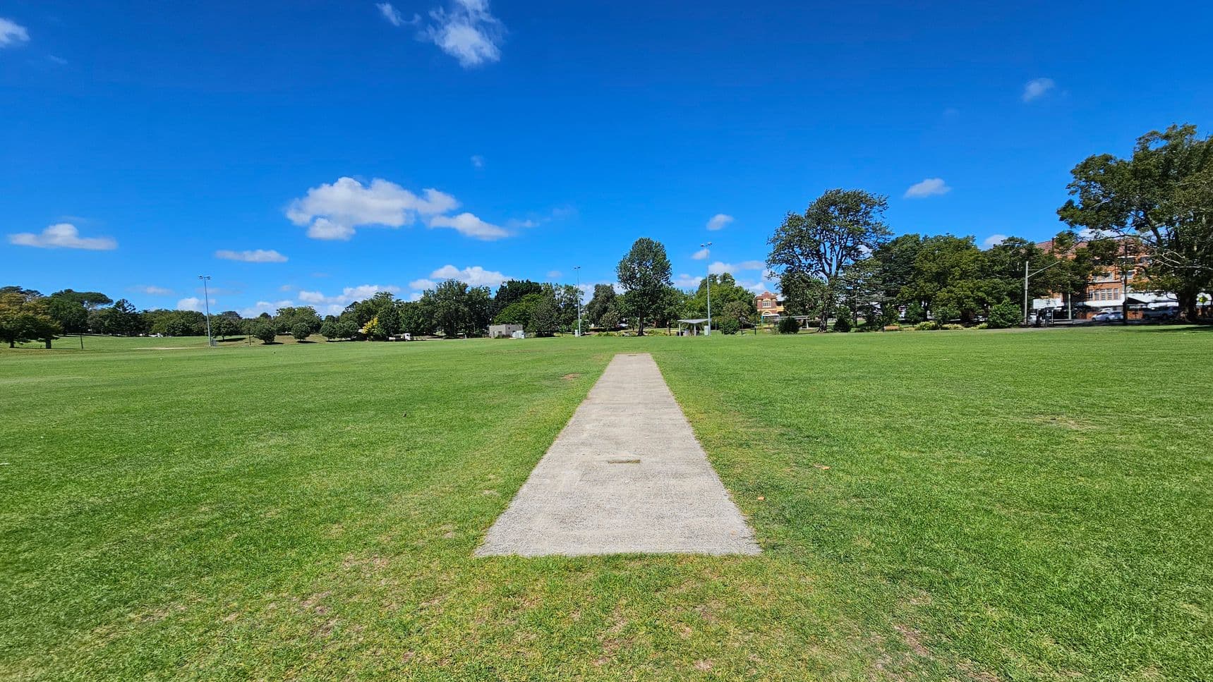 A cricket pitch is centred in a large, open grassy field. The pitch is a rectangular strip of lighter-coloured ground. Surrounding the field are trees and some buildings in the distance. There are a few light poles scattered around the field. The sky is clear with a few clouds.