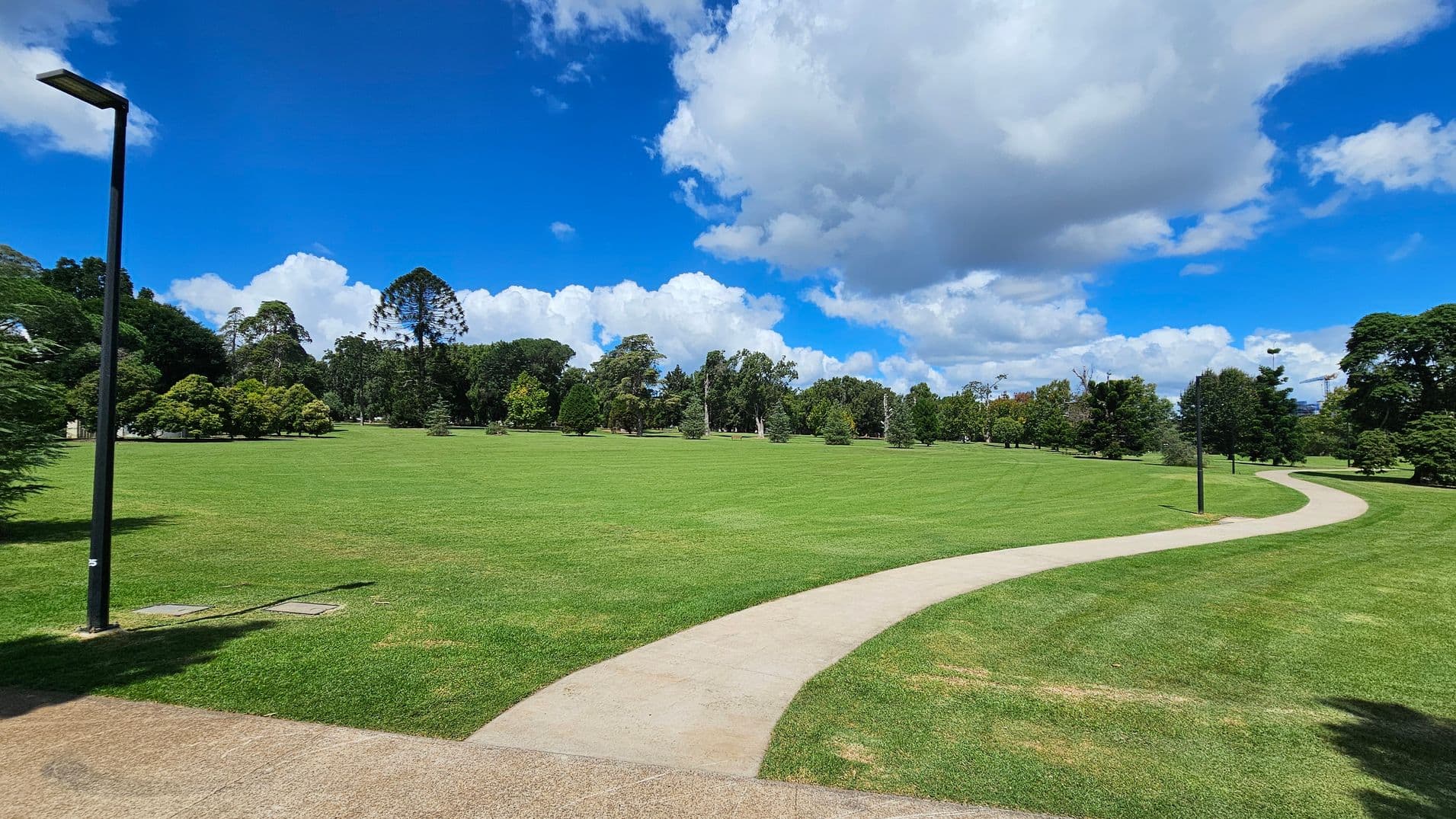 A wide, open grassy field with a winding concrete path running through it. On the left, a tall streetlamp stands near the path. The background features a variety of trees, including some tall and conical ones, under a sky filled with large, fluffy clouds. The grass is well-maintained, with patches of different shades of green.