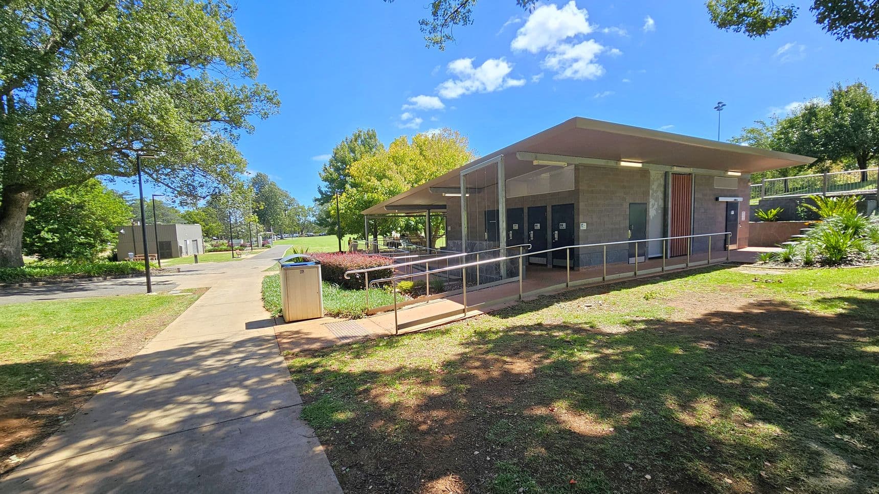 A public park scene with a modern restroom facility on the right, featuring a flat roof and a ramp for accessibility. The building is surrounded by small plants and a grassy area. A concrete path leads to the building, with a bin placed nearby. On the left, a large tree provides shade, and a lamppost is visible. In the background, there are more trees and an open grassy field. The sky is clear with a few clouds.
