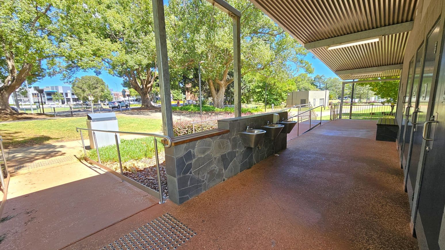 A covered outdoor area with a stone wall featuring two metal sinks on the right. The floor is a textured reddish-brown surface. Metal railings run along a ramp leading to a grassy area with large trees. In the background, there are buildings and a road with cars. The ceiling is corrugated metal with a fluorescent light fixture.