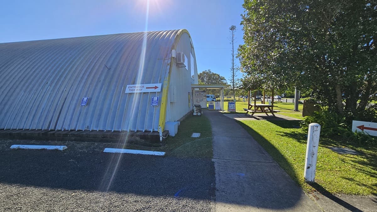A closer view reveals a large, dome-shaped metallic building with a corrugated surface to the left. To the right, there's a grassy area with trees, small signposts, and a picnic table. In the foreground, a concrete path runs centrally, leading to a gravel driveway. The vicinity of the building is covered with grass and gravel, and trees are also visible. Directional signage is affixed to the side of the building.