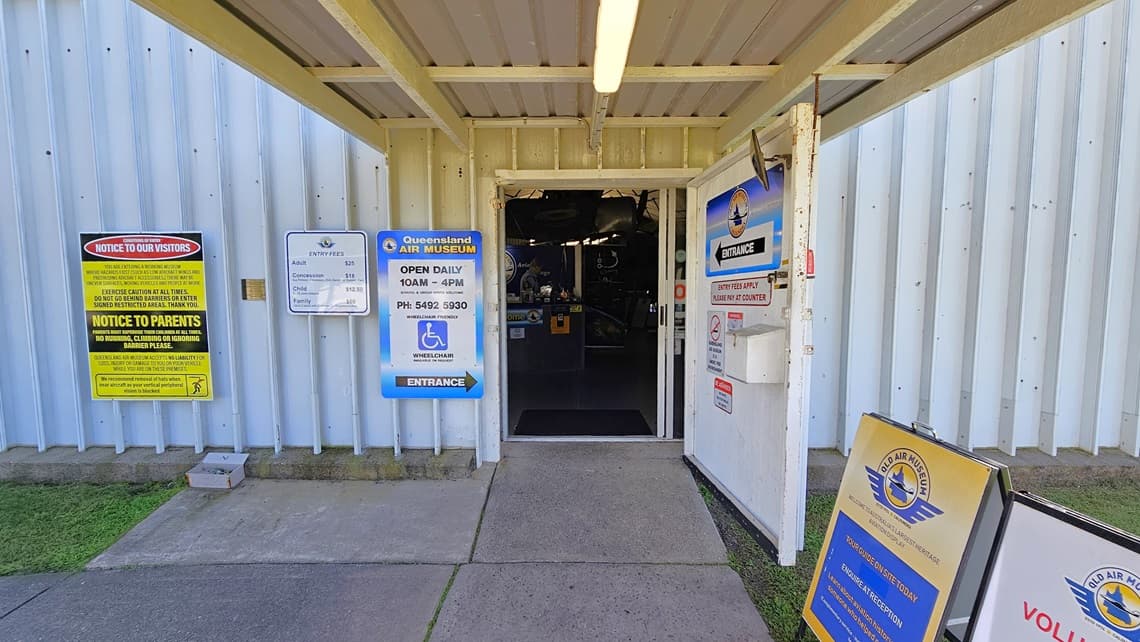 A corrugated metal wall displays several signs, including yellow notices for visitors and parents, and a blue one detailing the Queensland Air Museum's opening hours and wheelchair access. In the centre, an open door reveals additional signage, including a yellow and blue sign for volunteers, and offers a view into the building. The floor is concrete, and the ceiling features exposed beams with fluorescent lights.