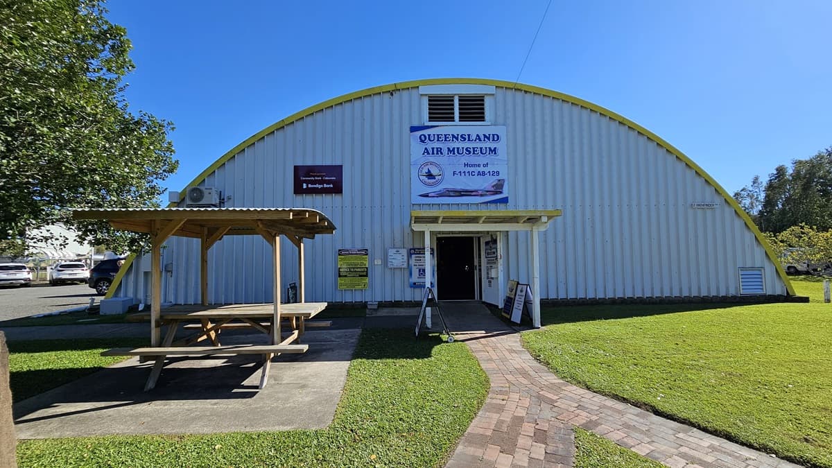 On the left, a picnic table sits under a corrugated roof. Next to it is a white corrugated metal building with a domed roof and a corrugated facade. Above the entrance, a sign reads "Queensland Air Museum." A brick path leads from the grassy foreground to the entrance, where two additional signs are located. Both the building and the gazebo are situated on a grassy area, with trees visible to the far left.