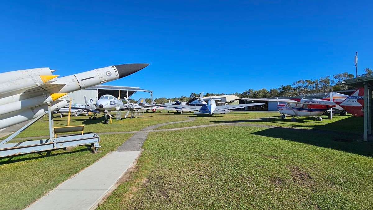 On the left, there is a missile displayed on a stand with various older aircraft positioned behind it. On the right, several small aircrafts are parked in a row, including a red and white aeroplane. In the background, additional aircraft are visible alongside a few hangars and some trees. The foreground features grass with a concrete pathway running through it.
