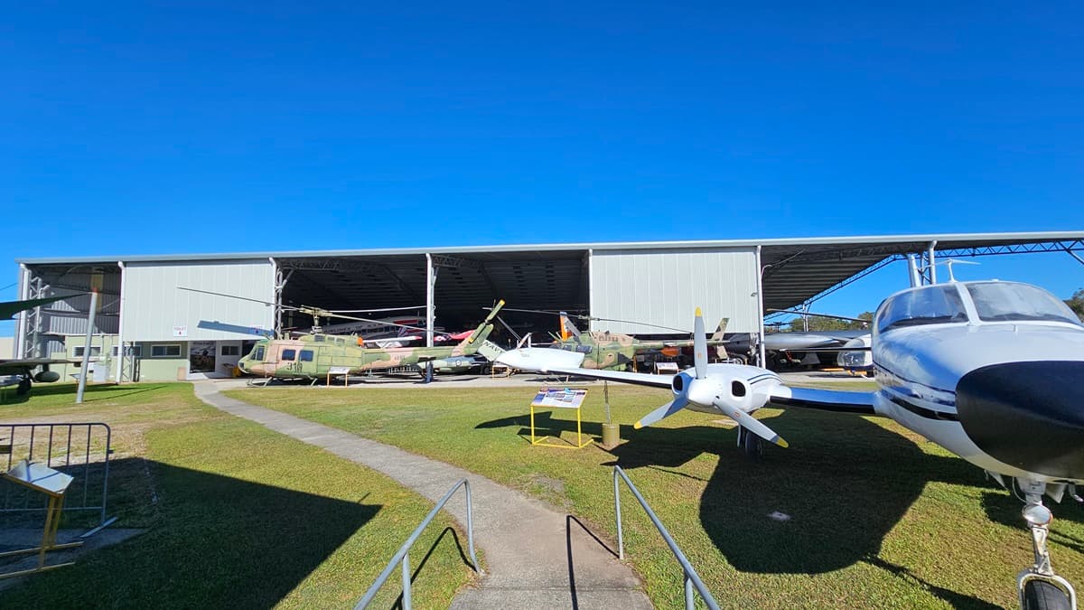 Several aircraft are arranged outdoors, with a large hangar extending horizontally across the background. On the left, various military-style helicopters and planes are positioned, with a paved walkway edged by grass leading to the hangar. On the right, the front of a white propeller airplane is visible, including the cockpit windows. The foreground features a well-kept grass lawn with shadows cast by the aircraft.