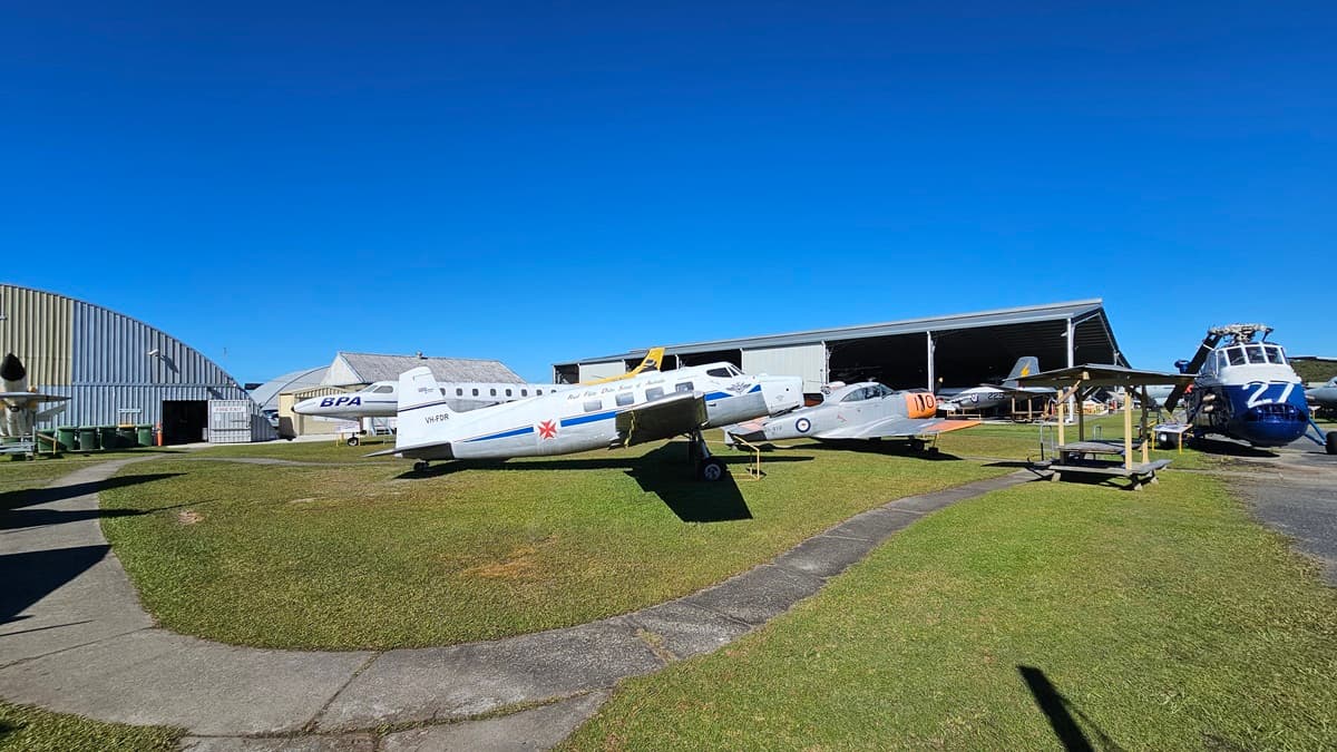 On the left, a silver and white aircraft with logos and registration numbers is parked on grass, with a large arched hangar in the background and several smaller buildings nearby. On the right, another aircraft with blue and white markings is partially inside an open hangar, alongside a blue and white helicopter. The foreground features a concrete pathway curving through the green grass.