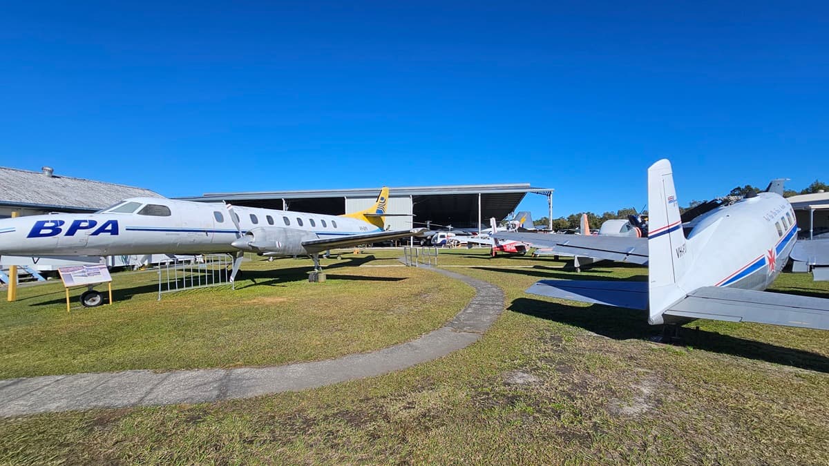 On the left, there is a white aircraft with "BPA" written on the side, its wings stretching out. On the right, another aircraft, predominantly white with blue and red stripes and a visible tail fin, is parked. The ground is covered with grass, and a narrow concrete path winds through the area. In the background, several more aircraft are parked, and a large hangar with an open front is visible, housing additional planes.