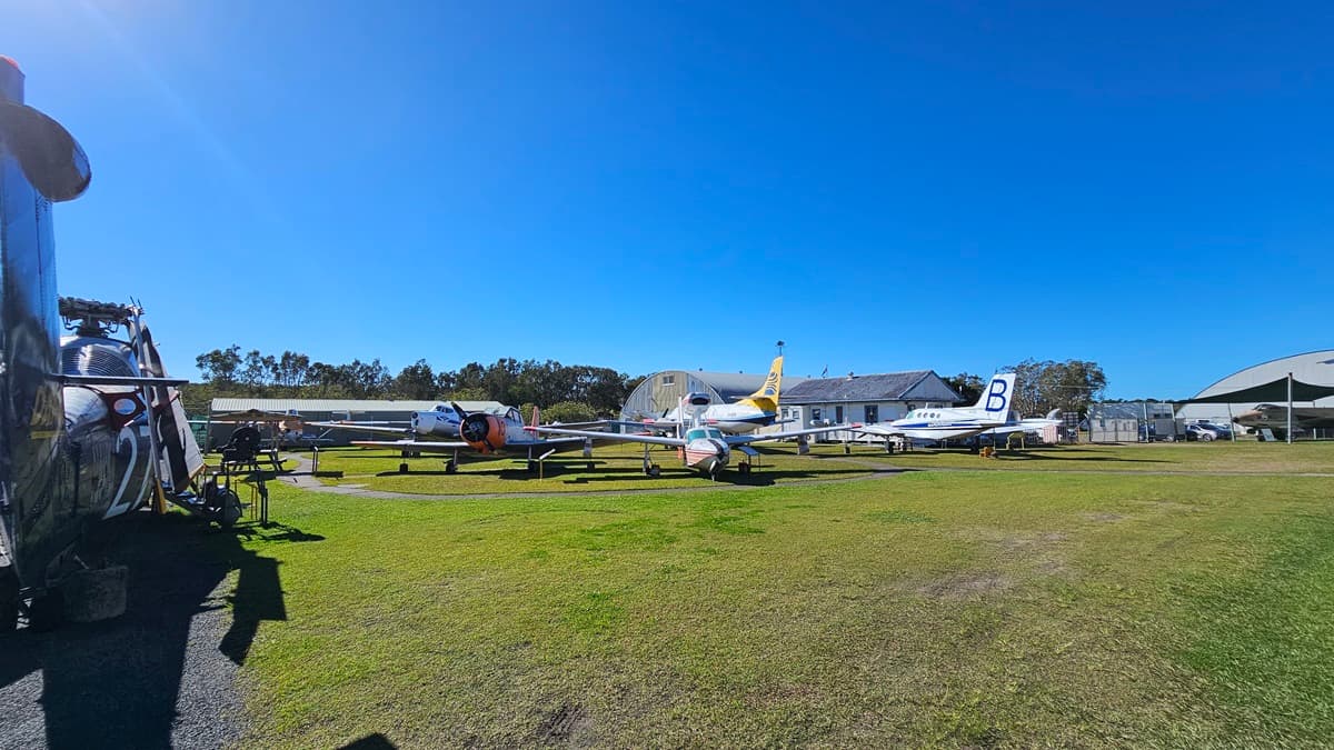 In the middle, several vintage aircraft are parked on a grassy field, each with distinct markings and colours. One has a prominent yellow nose and another features a large "B" on its tail. To the right, a shed and additional aircraft are visible, including one with a silver body. Trees line the background, and the grass underneath is green and well-kept, with patches of bare earth.