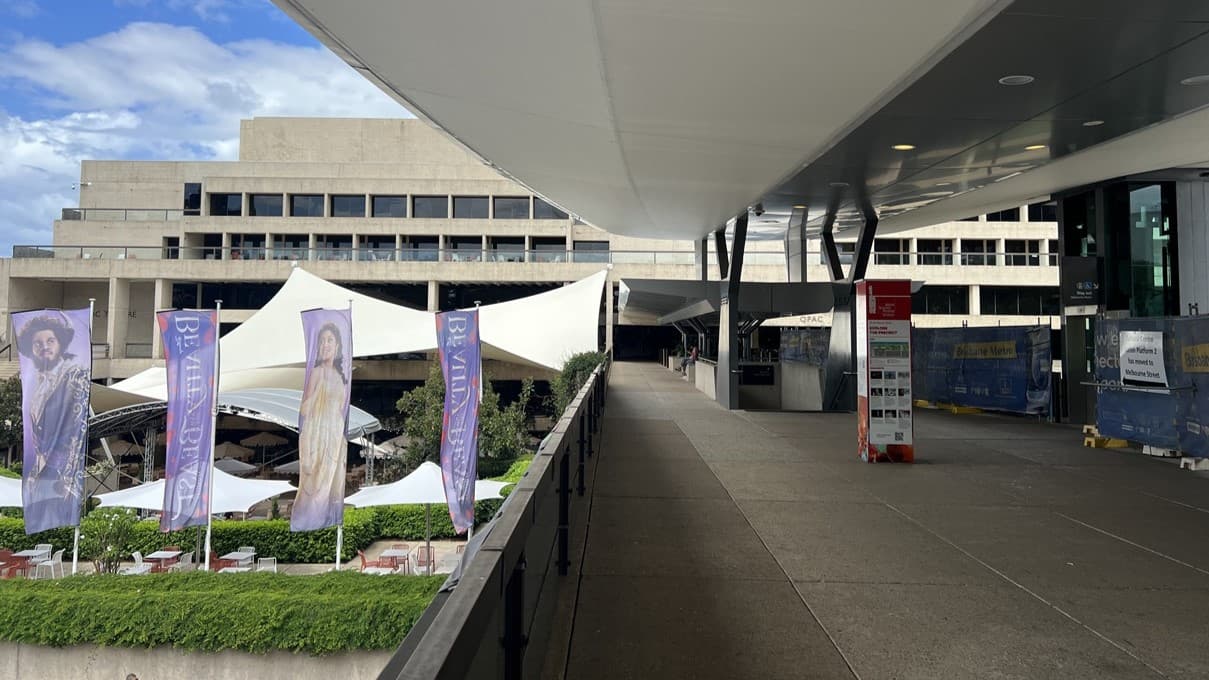 To the left, a row of violet banners line a footpath, with greenery and grass on either side. Tables and chairs are neatly set beneath sizable umbrellas. On the right, a corridor stretches beneath a broad white canopy, guiding towards a multi-storey building. Stairs with handrails descend nearby. Banners and construction barriers are seen on the right side.
