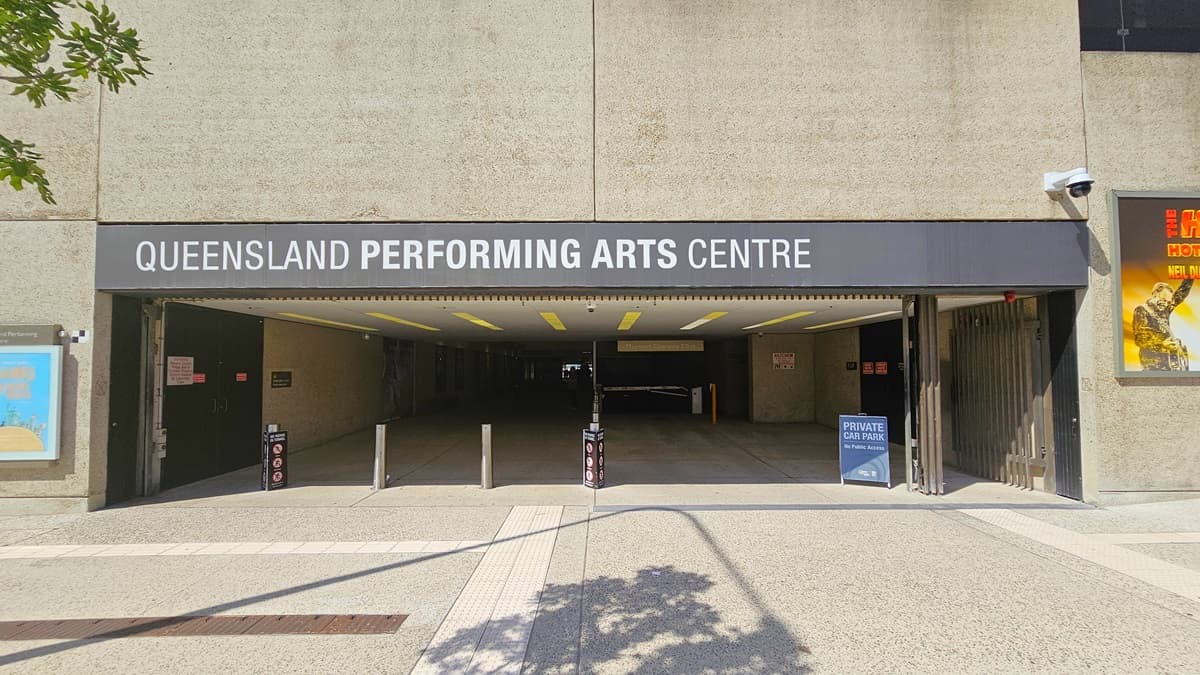 An underground car park, with a low ceiling and dim lighting, has the facility name mounted at the front. Bollards accompanied by warning signs border the left side. A sign stating "Private Car Park" is positioned on the right, with additional signage affixed to the interior walls. Large posters adorn both walls. The building's façade showcases a concrete design, and the floor, featuring truncated domes, has a finished surface.