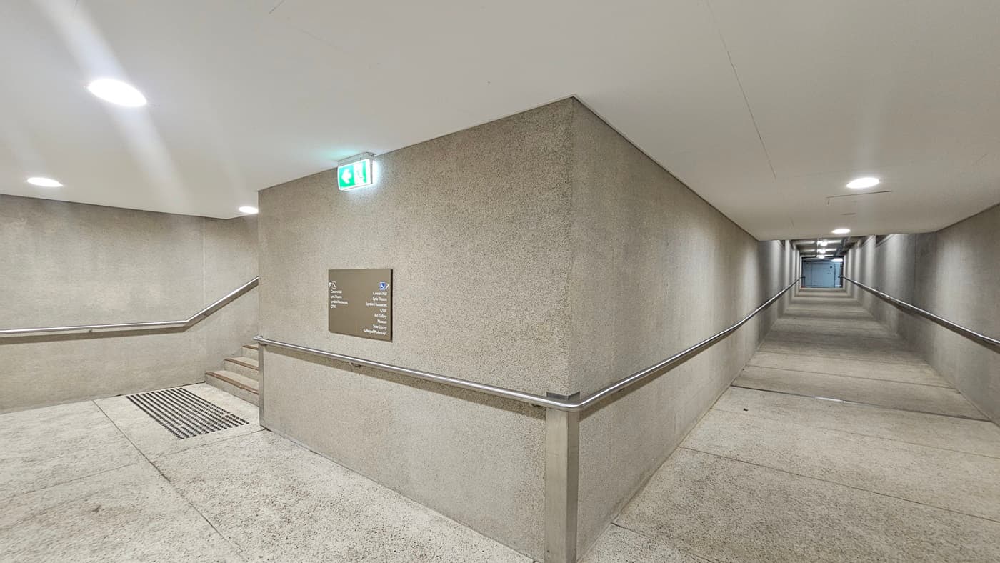 On the right, there is a long pedestrian ramp equipped with handrails. On the left, there are stairs with handrails and truncated domes are installed before the stairs. A directional signage is affixed to the middle wall, along with an exit sign. The walls and floors are made of concrete, and the ceiling is white with LED lights.