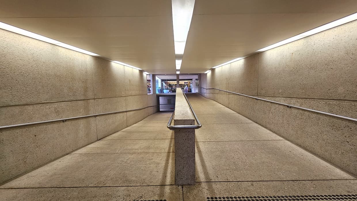 A winding ramp with handrails and truncated domes for safety. A concrete partition secures the ramp. Concrete floor and wall, white ceiling with fluorescent lights. At the end of the ramp, retail shops are visible.