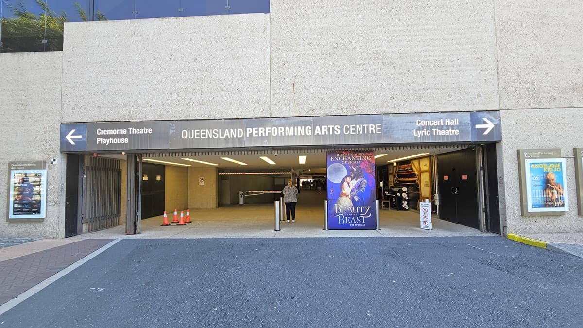 The tunnel entrance with facility signage indicating Queensland Performing Arts Centre and directional arrows to other theatres. Posters decorate the walls. Colourful posters and bollards line the front, alongside orange safety cones and other signs. Inside, retail shops are visible. The building's front has a concrete facade.