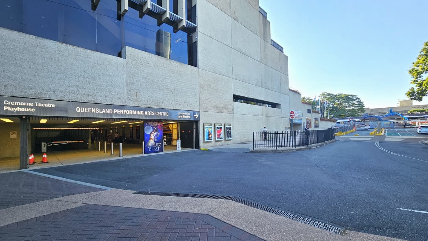 At the tunnel entrance, facility signage directs to Queensland Performing Arts Centre and other theatres. Posters adorn walls. Colourful posters and bollards line the front, with orange safety cones and signs. The building has a concrete and glass facade. To the right, a two-way road with metal fencing and barricaded section for construction.