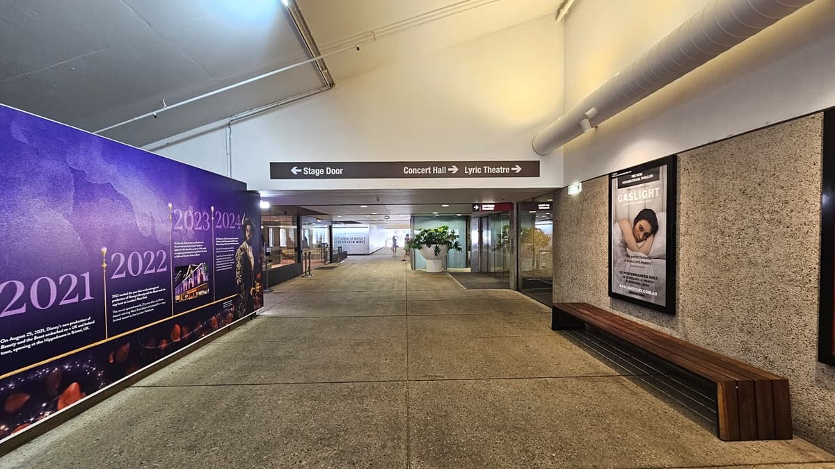 The pedestrian tunnel has a white pitched ceiling with sizable aircon vents on the right. Below, a wooden bench chair. A large poster fills the left wall. Further in, retail shops visible, with indoor plants in a large white pot. Directional signage also present.