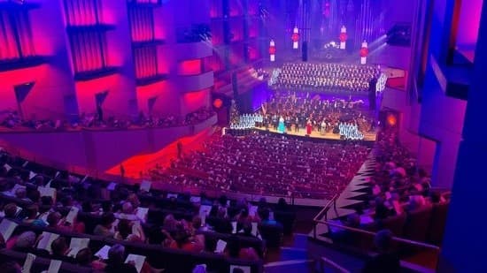 A panoramic view of the concert hall, including the stage, occupied by spectators and performers. The area is dimly lit with lights illuminated from the concert stage. Chairs are arranged in rows, stalls, and boxes. Light fixtures hang from the high ceiling. The wall is adorned with timber decorations.