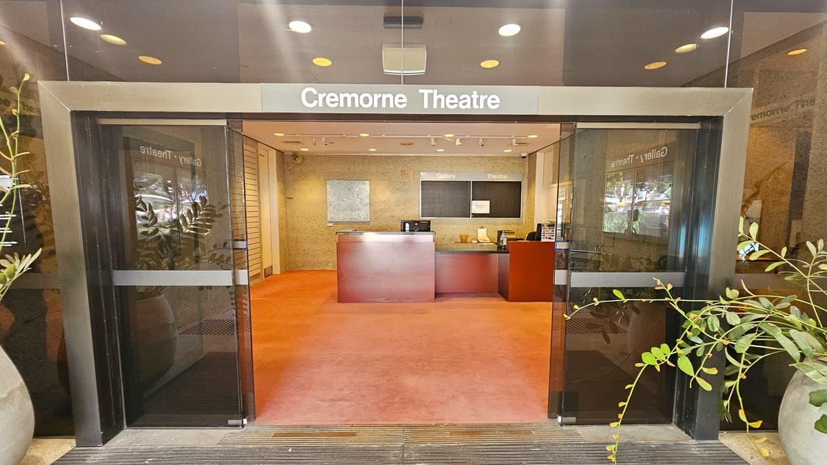 The entrance features an automatic glass sliding door. a signage that reads "Cremorne Theatre" is placed above. Inside, a reception desk with a brown facade holds computers and posters. Behind the desk, an abstract painting hangs on the cream wall. The floor is covered in pink carpet, and the ceiling features white light fixtures. Large potted plants adorn the entrance.