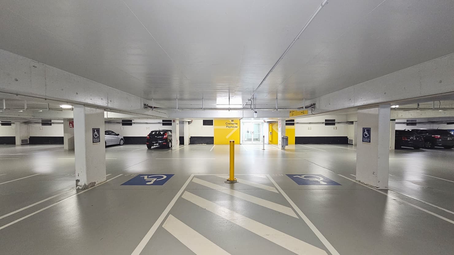 The accessible parking area is marked with designated accessibility symbols, accompanied by a yellow bollard in the centre. The ceiling is low and painted white, with a visible glass door entrance at the far end. Yellow directional signage is positioned on the middle wall, while vehicles are parked on the opposite side.