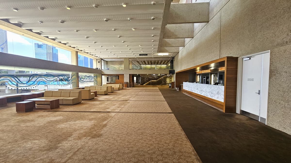 The lobby features a long front desk with a wooden and marble façade against a beige wall. Various drinks are displayed in the fridge. Adjacent is a white door. The carpeted floor extends towards the seating area with sofas. The left wall features a glass-wavy accent of blue and white. The ceiling is lined with parallel lighting fixtures and aircon vents.