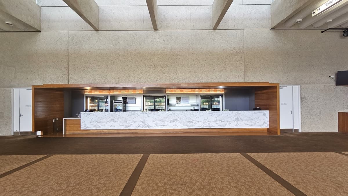 The lobby showcases a long front desk with a wooden and marble façade against a beige wall. Various drinks are displayed in the fridge. Adjacent is a white door. The floor has printed brown carpet, and the ceiling is lined with parallel lighting fixtures and aircon vents.
