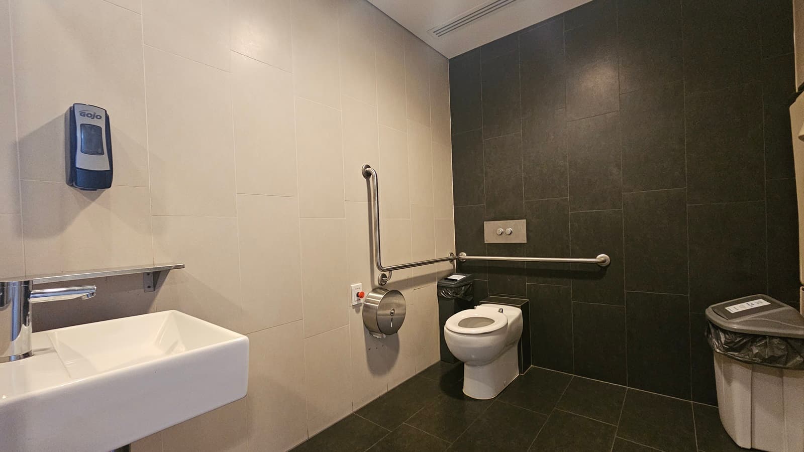 The accessible bathroom features a toilet bowl with a hand grab positioned on the corner, along with a tissue dispenser and rubbish bin. Another rubbish bin is set against the wall on the right. A reminder is affixed to the wall, and a red push button to lock the door is also visible. A wash basin is mounted to the wall on the left along with a liquid soap dispenser. The floor has grey tiles, and the ceiling is white. The wall combines black and cream tiles.