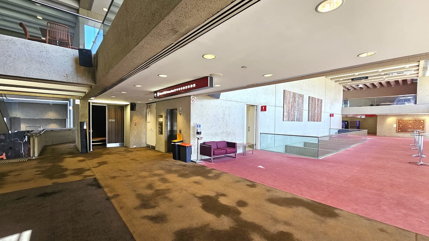 An interior space with a carpeted floor; on the right, there's toilet with signage above. Posters are affixed to the cream walls. The front is enclosed with glass railings with seating. A cushioned chair is positioned near the toilet door along with a sanitising machine. On the left, a ramp with railings leading up, a lift, and a rubbish bin nearby. The carpet transitions from pink to brown and the ceiling is white with LED lights.