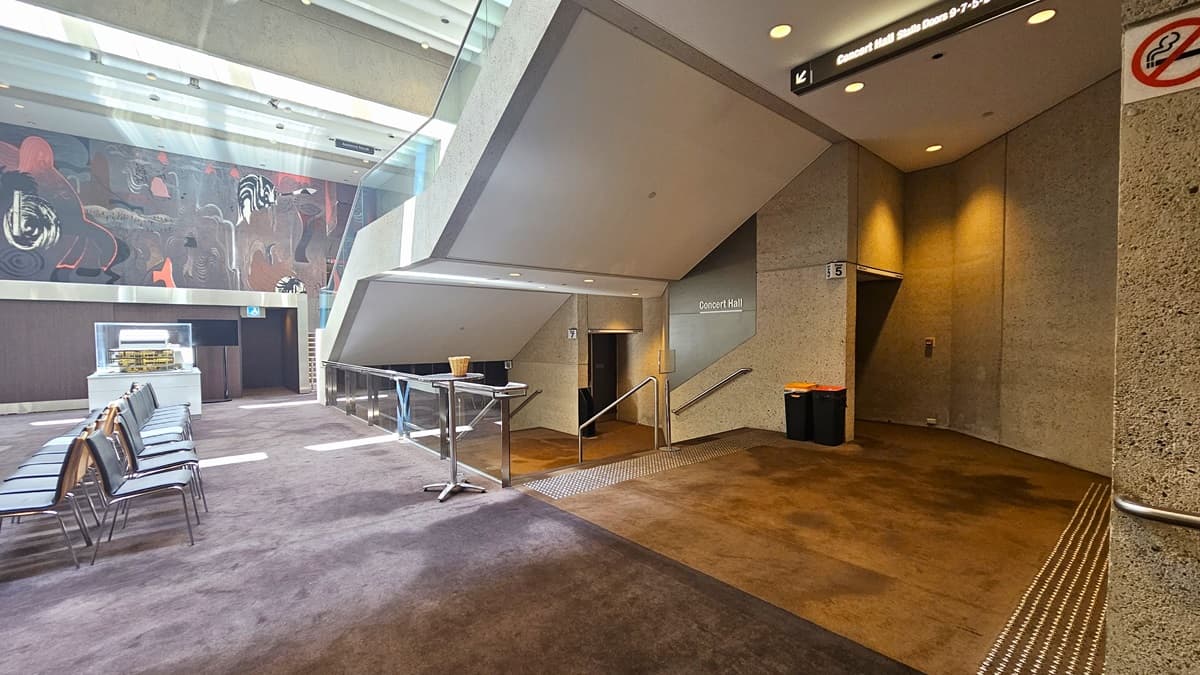 To the right, there is a staircase with handrails equipped with truncated domes, while a large mural decorates the wall. Underneath, there is another set of staircases with truncated domes and handrails leading to the Concert Hall. Signages are affixed to the walls, and two rubbish bins are placed beside them. The floor is carpeted, and the ceiling is white.
