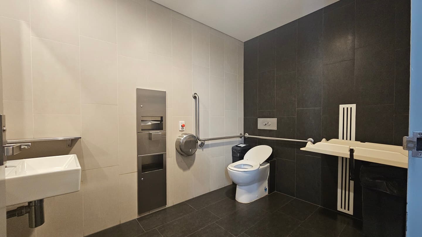 The accessible bathroom features a toilet bowl with hand grabs on the corner and tissue dispensers mounted to the wall. A change table is positioned to the right, with a rubbish bin underneath. To the left is a wash basin. The wall has black and white tiles, and the floor is black.