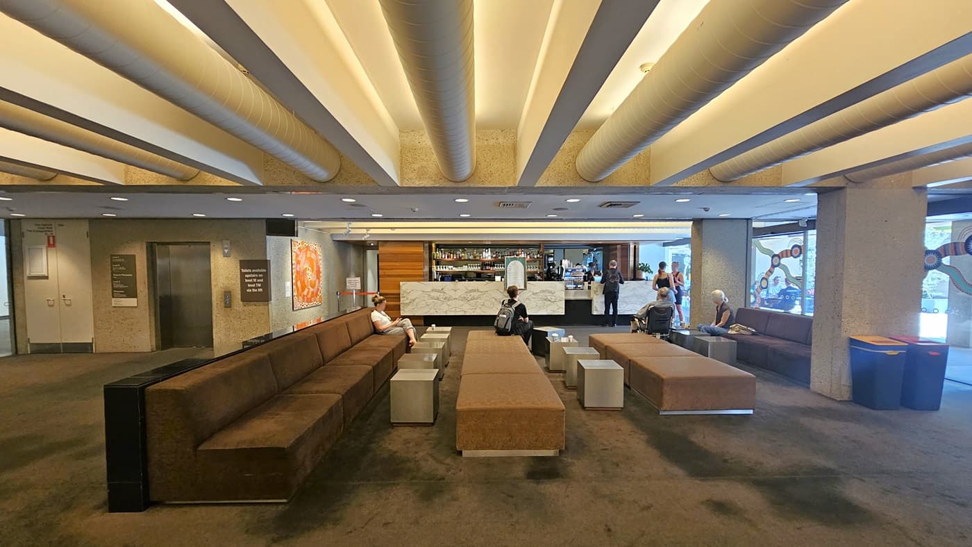 To the right, a sitting area furnished with lounge chairs and coffee tables. Adjacent, a bar counter displays wines, liquors, and drinks, featuring a marble design front. A lift positioned to the left side, equipped with directional signages. A large painting hangs on the middle wall. The floor has dark grey carpet, and the ceiling features white with large aircon vents and LED lights. Guests are visible in the area.