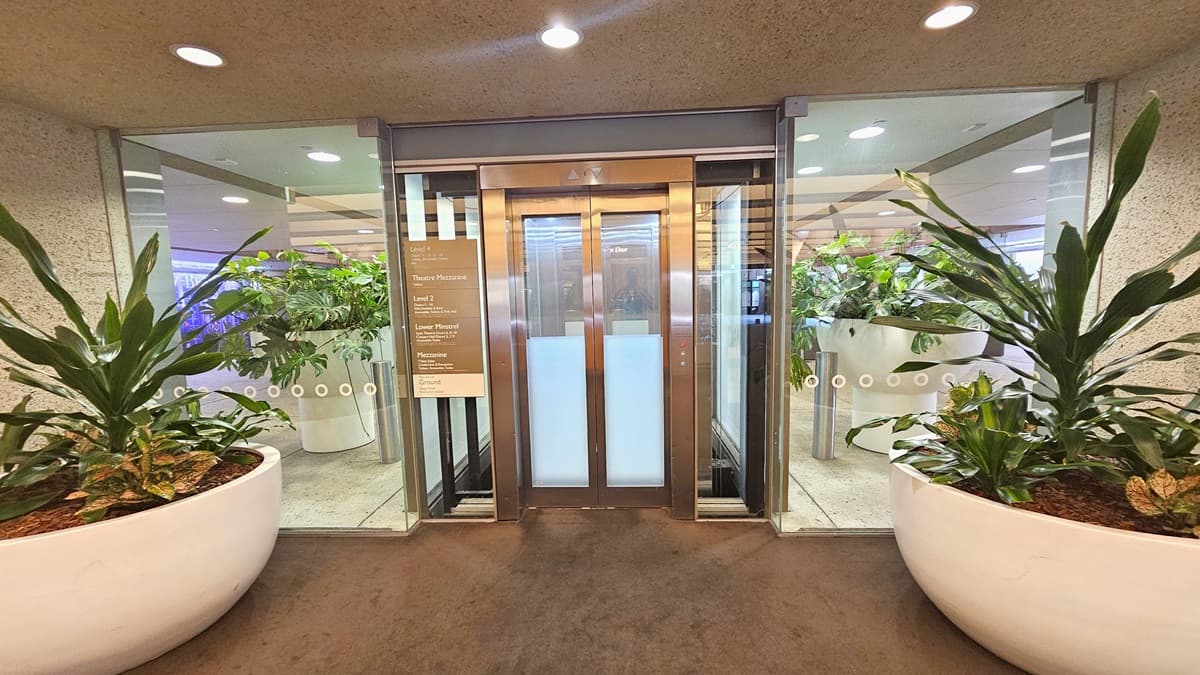 In the middle, a lift equipped with a poster. Large pots with indoor plants on both sides and two sets of plants inside. The ceiling and wall are concrete with LED lights, and the floor has dark grey carpet.