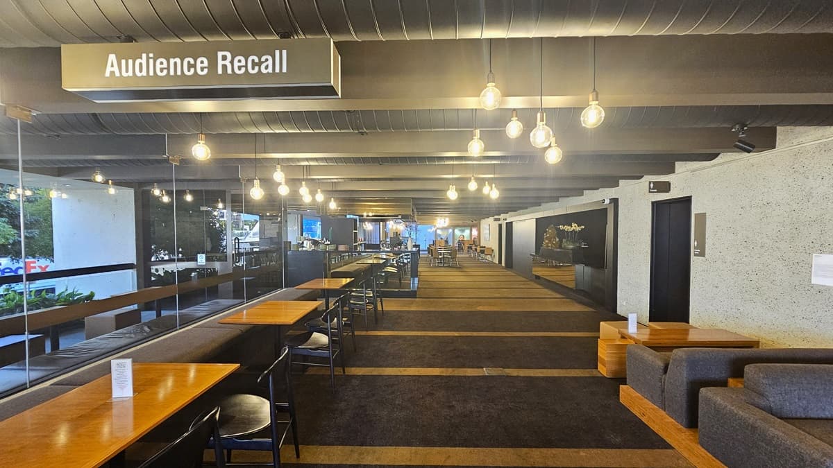 A carpeted area with rows of tables and movable chairs, along with cushioned bench chairs positioned on the left. On the right, couches and coffee tables are arranged against the wall. Further inside, more seating. Brown carpeted floor, with a dotted white wall on the right and a glass wall on the left. The ceiling features large aircon vents and light fixtures. A sign reading "Audience Recall" hangs from the ceiling.