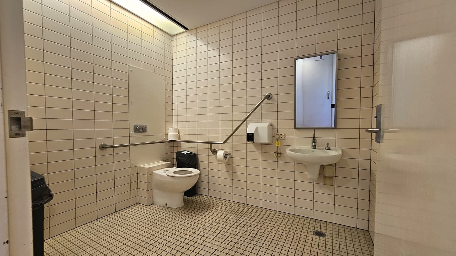 Within the accessible bathroom, a toilet bowl with handrails is situated in the corner. Beside it, a tissue dispenser and a rubbish bin are placed. Adjacent to the toilet, there is a wash basin and a wall-mounted liquid soap dispenser. Another rubbish bin is positioned on the left, near the wall. The walls and floor are adorned with white tiles, while the ceiling also maintains a white colour scheme.