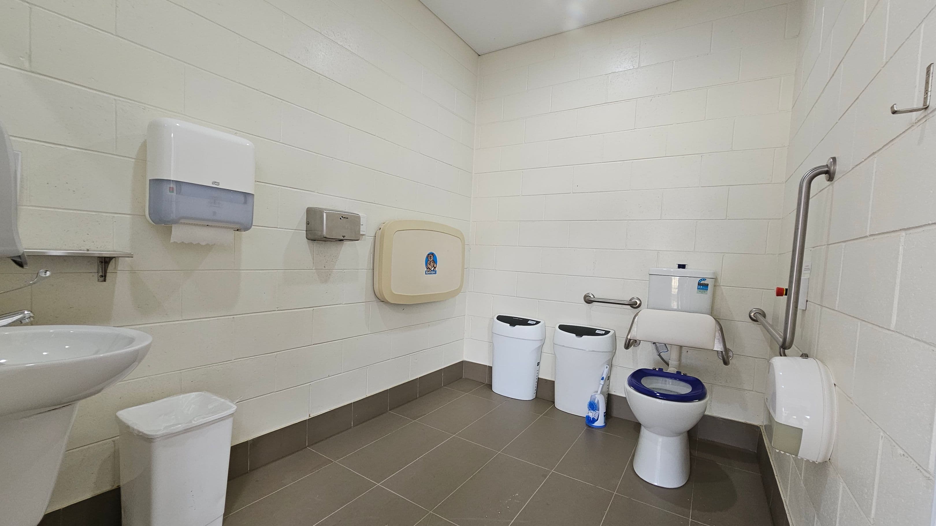 A public restroom with white tiled walls and a brown-tiled floor. On the left, there is a sink with a white bin beneath it, and a paper towel dispenser above. In the centre, a baby changing station is mounted on the wall. To the right, there are two white bins, a toilet with a blue seat, and a toilet brush. Stainless steel grab bars are installed around the toilet, and a toilet paper dispenser is mounted on the wall. The ceiling is plain and light-coloured.