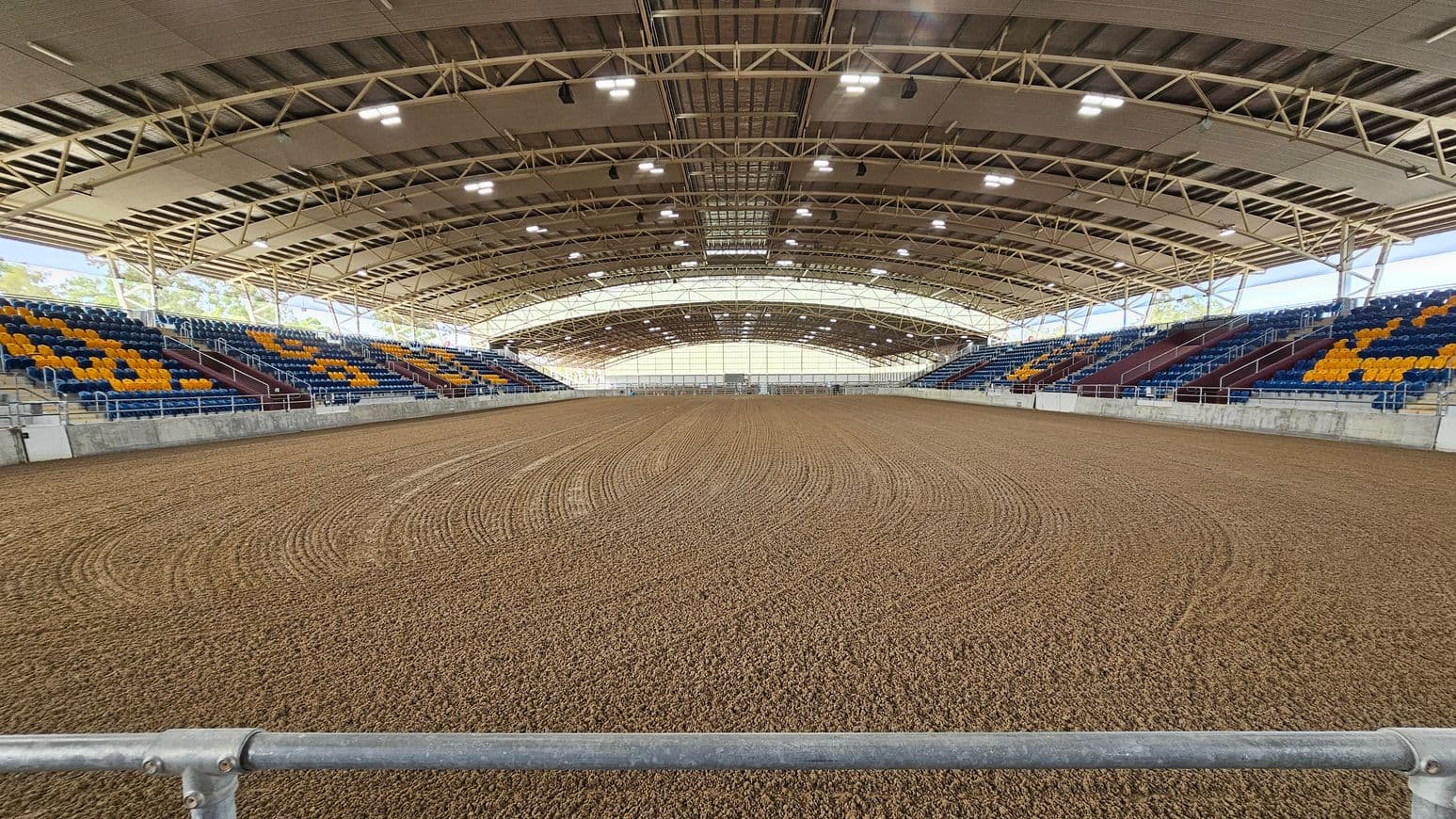 An indoor arena with a dirt floor featuring circular patterns. On both sides, there are tiered seating areas with blue and yellow seats. The ceiling is supported by a metal framework with multiple lights. The background shows a large, translucent wall allowing natural light to enter. A metal railing is visible in the foreground.