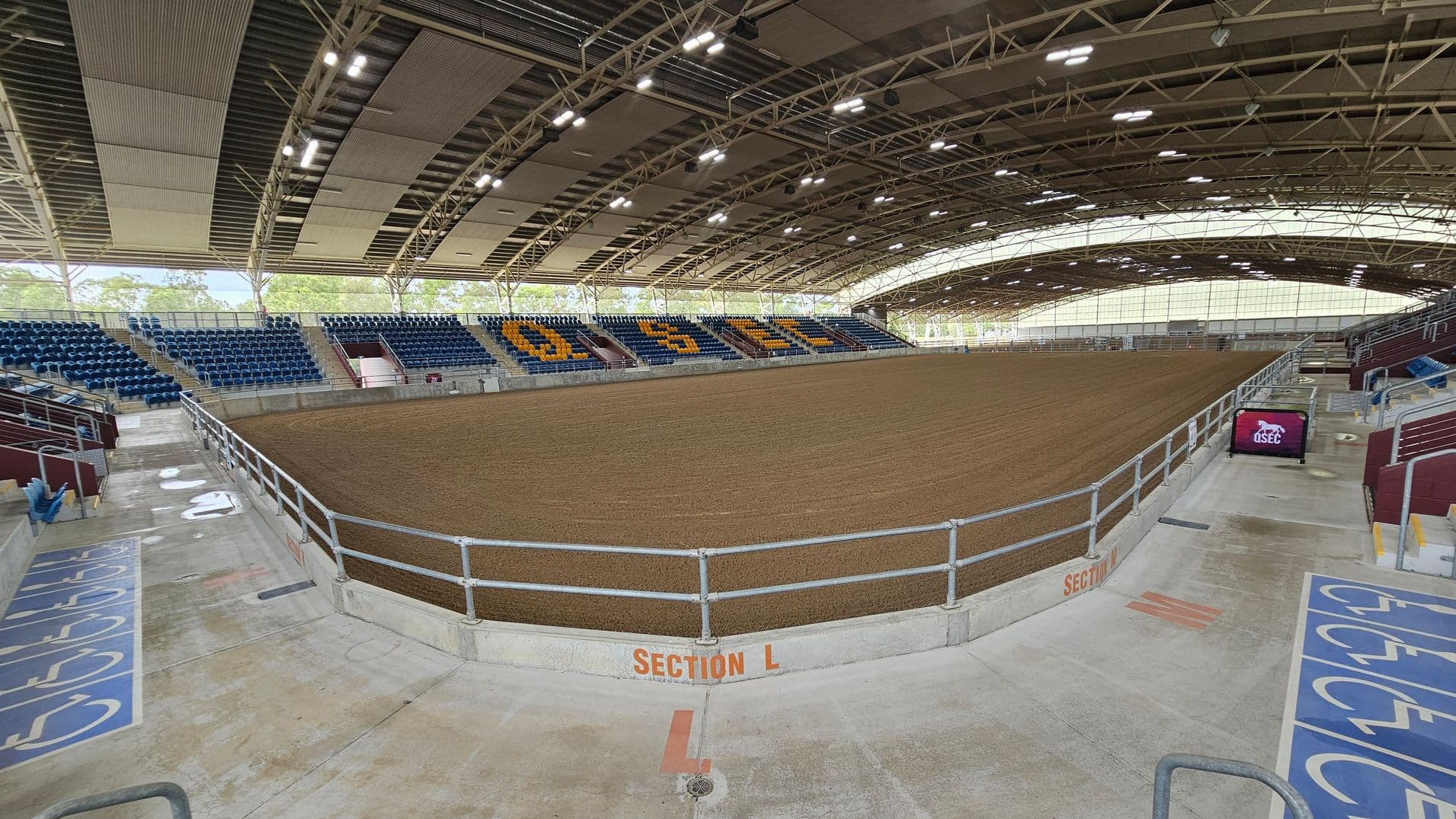 An indoor arena with a large dirt floor in the centre, surrounded by tiered seating. The seats on the left and right are blue, with some sections displaying yellow letters. The ceiling is arched with exposed beams and lighting fixtures. The floor is concrete with marked sections, including a designated area for accessibility. A railing separates the seating from the arena floor. A digital screen on the right displays a logo. The background shows trees visible through the open sides.