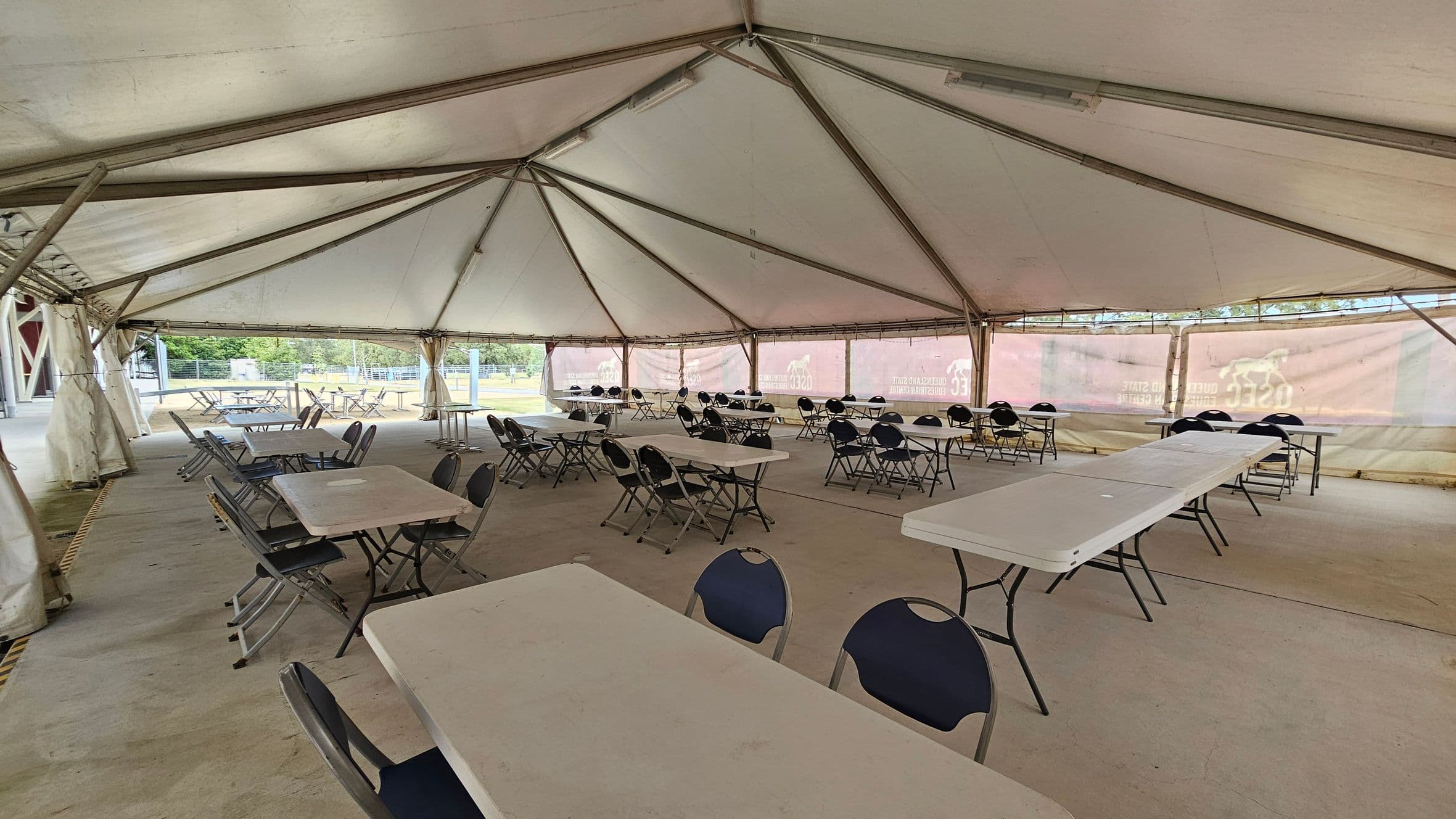 Inside a large tent with a high, triangular ceiling supported by metal beams. The floor is concrete. Several rectangular tables are arranged in rows, each surrounded by folding chairs. The tent walls are partially transparent, allowing a view of the outdoor area with greenery and structures in the background. The space appears set up for an event or gathering, with ample seating available.