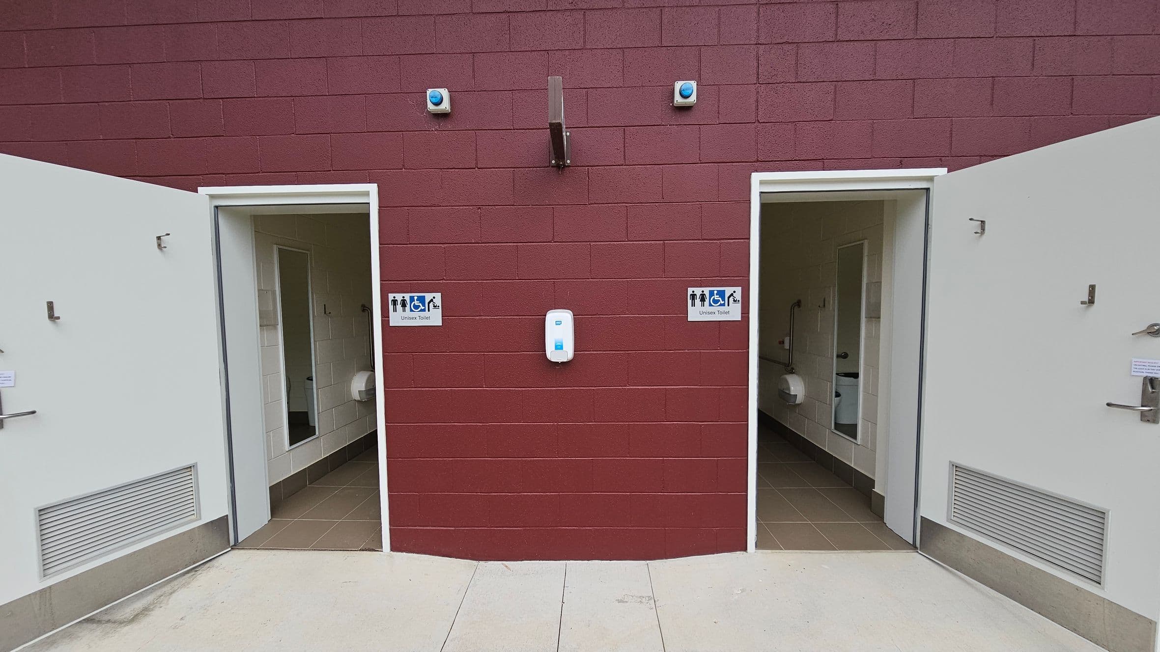 Two open doors on a maroon brick wall lead to unisex toilets, each marked with a sign showing accessibility symbols. Between the doors, a white dispenser is mounted on the wall. The interior of the toilets reveals white tiled walls and beige floors, with visible toilet paper dispensers. The floor outside is light grey concrete. Two small blue lights are above the doors, and a metal fixture is centred above the dispenser.