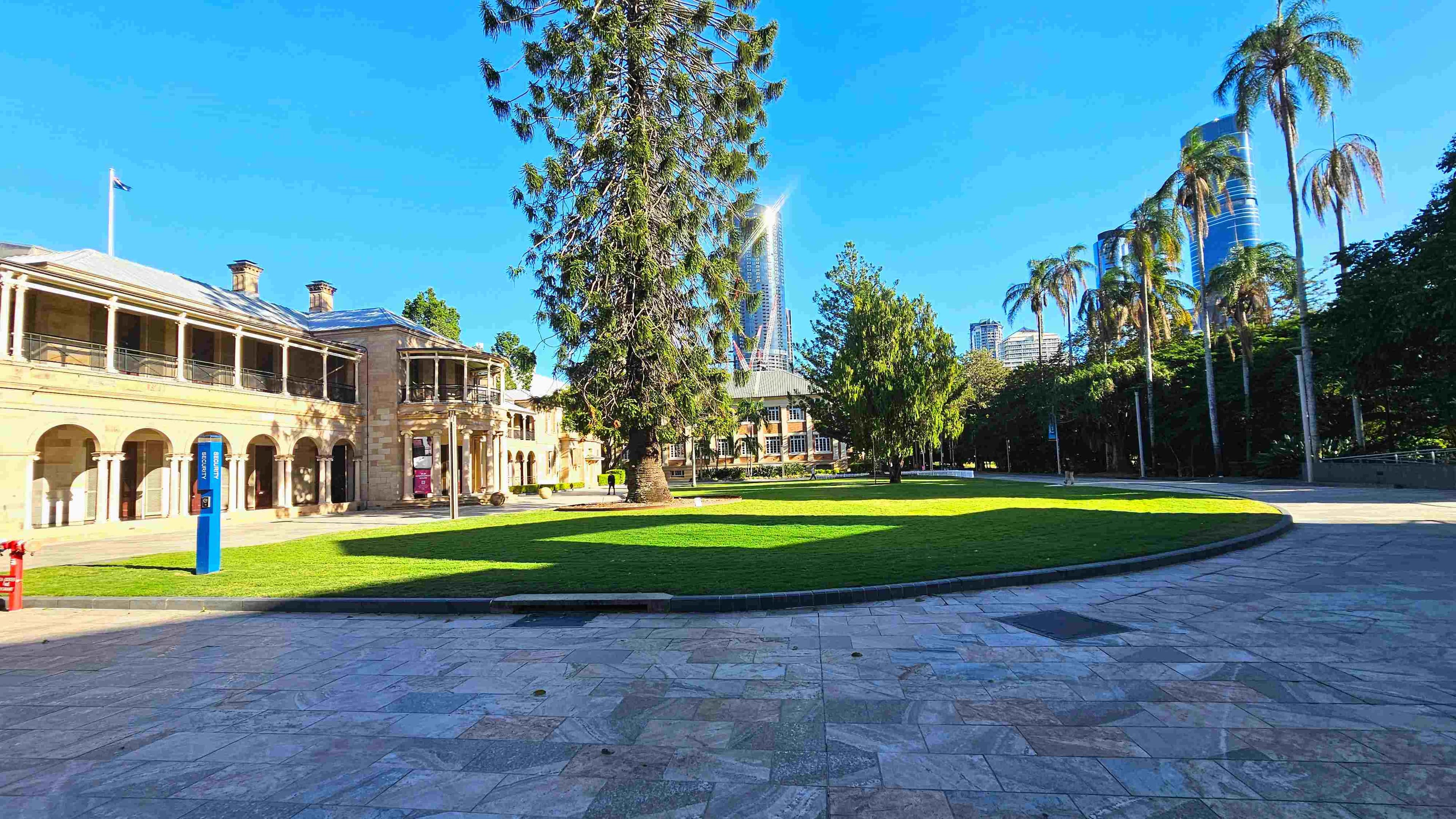 A spacious outdoor area with a large, well-maintained lawn bordered by a stone pathway. On the left, a historic building with arched windows and a flagpole stands prominently. Tall palm trees line the right side, and modern skyscrapers are visible in the background. A blue information sign is near the building, and the area feels open with a mix of natural and architectural elements.